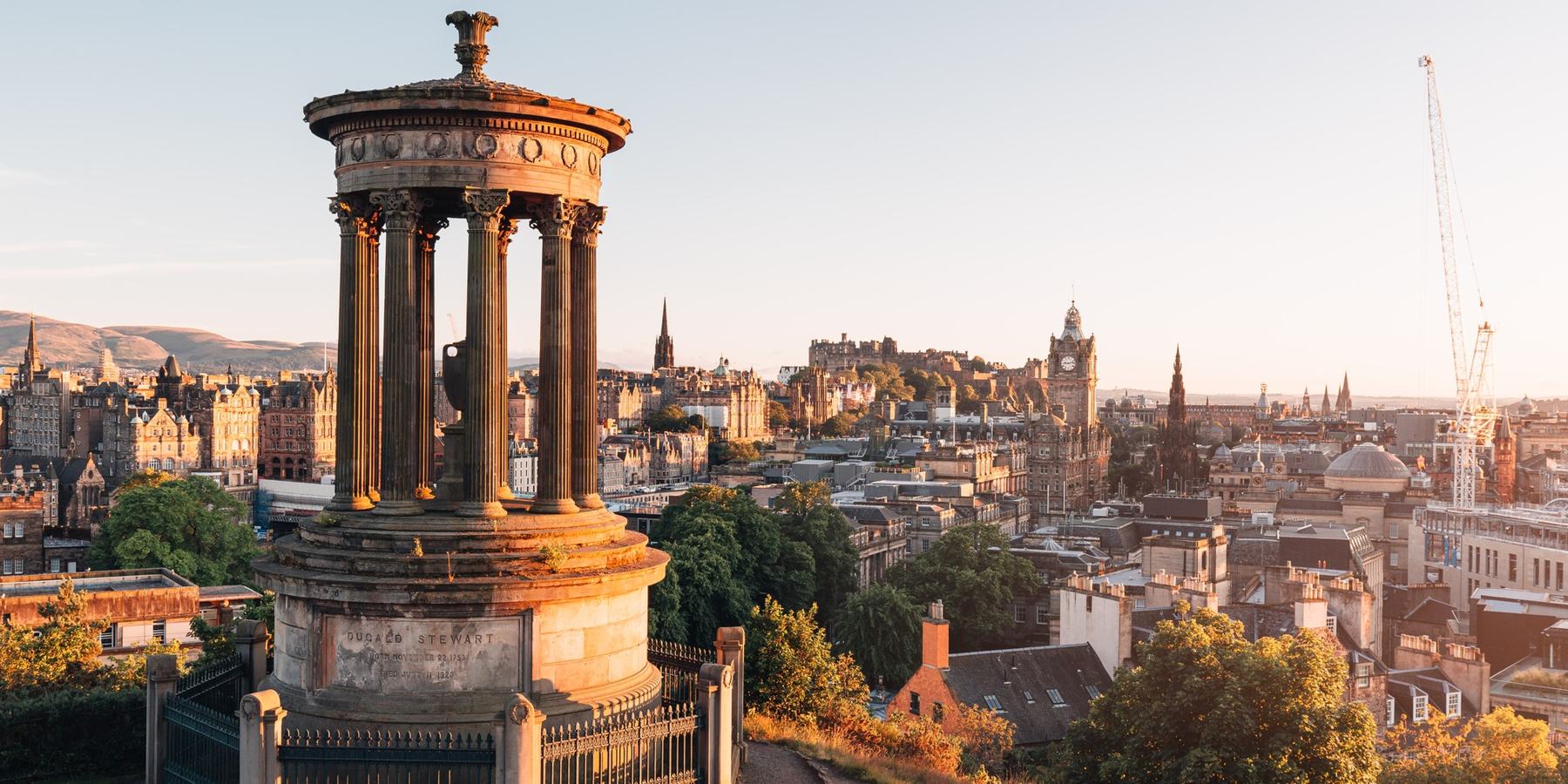 The Dugald Stewart Monument on Calton Hill at golden hour, with Edinburgh Castle, the Balmoral clock tower and the Scott Monument along the skyline behind it